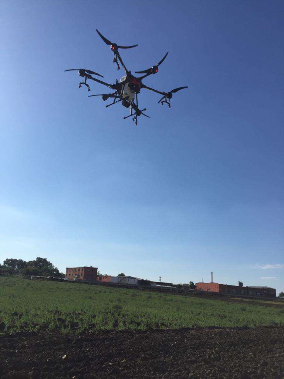 A six-rotor DJI Agras T-16 spraying drone in flight, seen from below against a clear sky, over a field with low brick buildings behind.