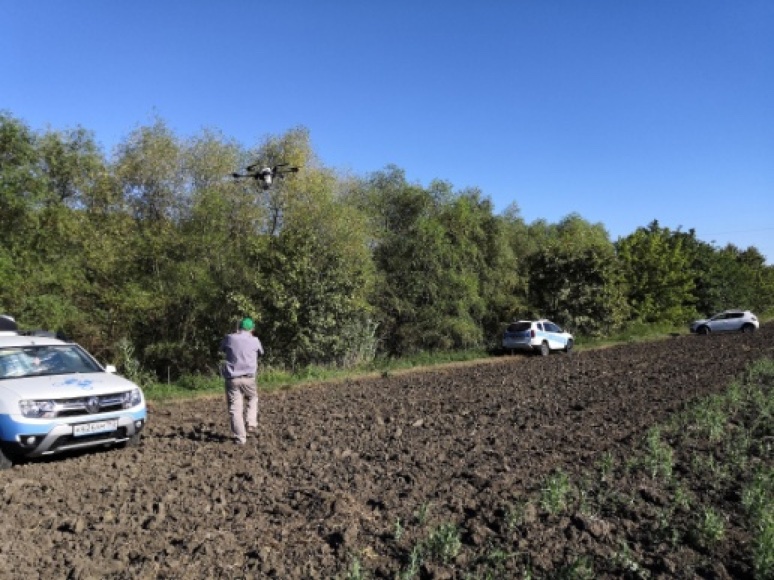 An operator at the edge of a ploughed field watches a drone above the tree line; cars are parked along the tree line.