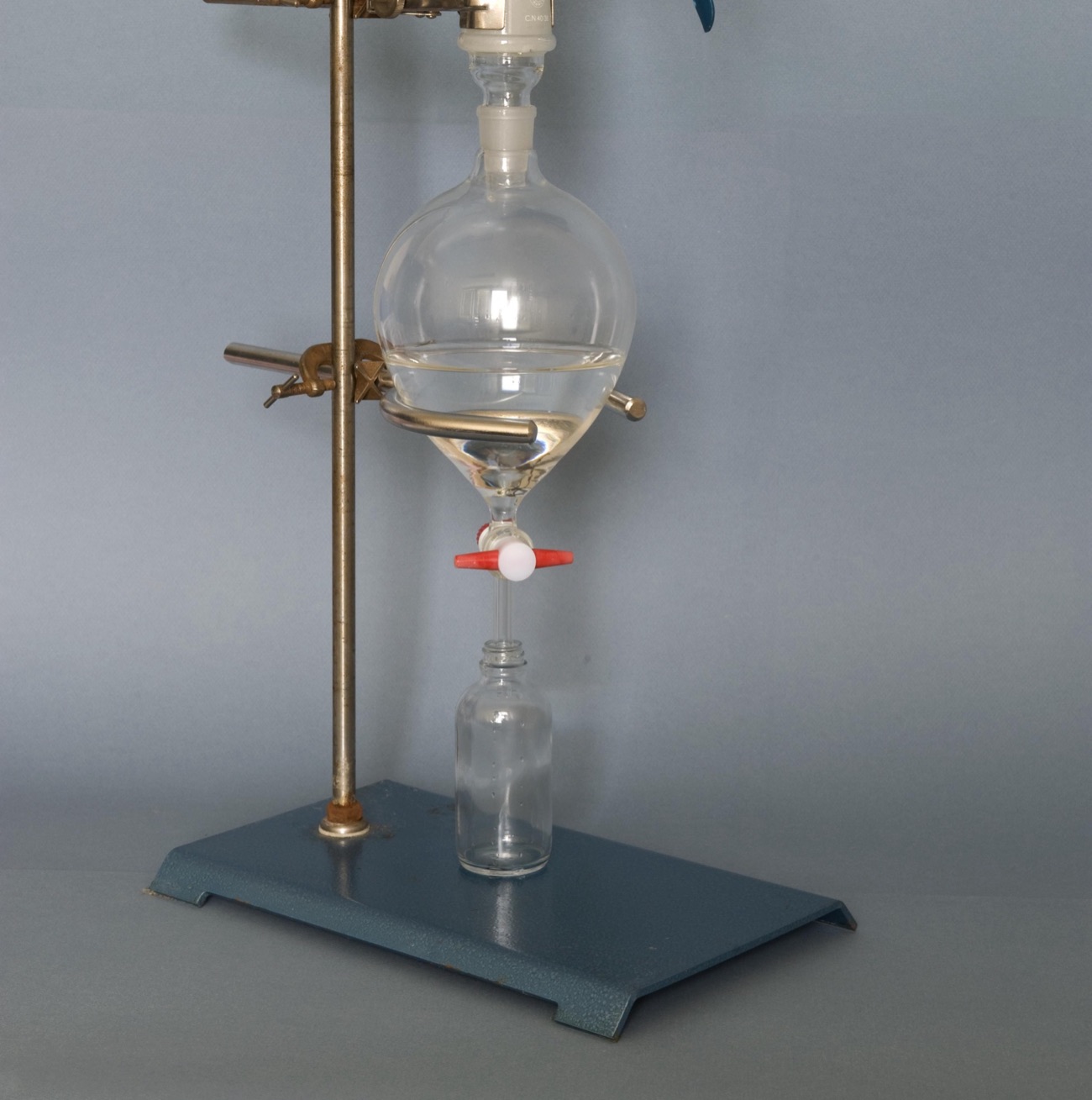 A glass separating funnel clamped to a laboratory stand, a clear liquid filling its lower half, its stopcock closed above a small empty bottle, on a grey background.